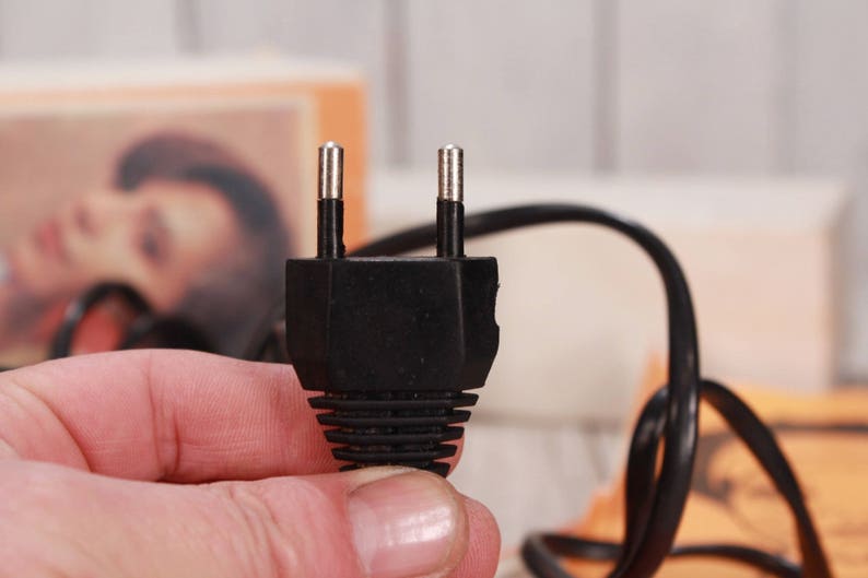 May include: A black electrical plug with two metal prongs. The plug is held in a hand, with a black cord extending from it. The background is blurred, showing a light-colored surface and a box.
