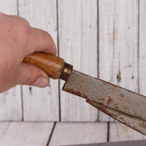 May include: A vintage woodworking tool with a wooden handle and a metal blade. The handle is light brown, and the blade shows signs of rust. The tool is held against a white wooden background.