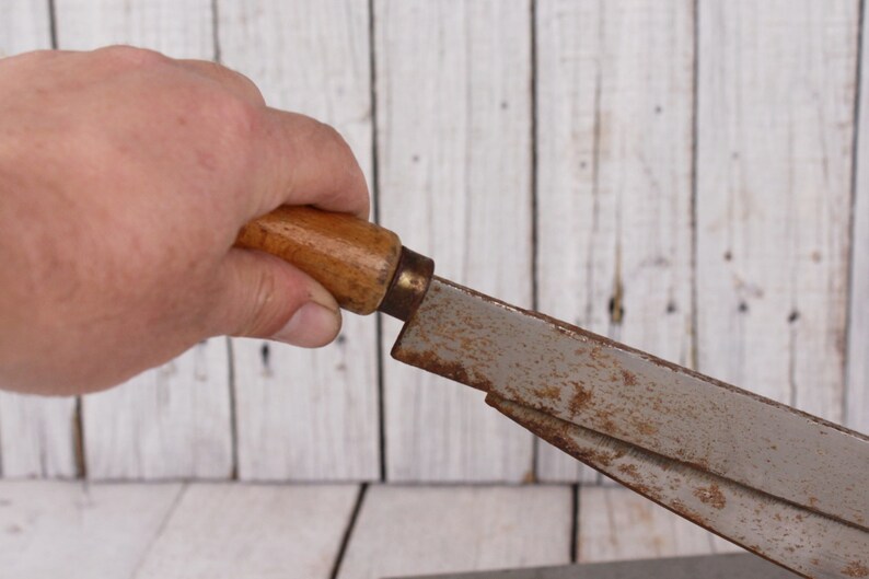 May include: A vintage woodworking tool with a wooden handle and a metal blade. The handle is light brown, and the blade shows signs of rust. The tool is held against a white wooden background.