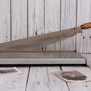 May include: Vintage metal paper cutter with a wooden handle. The blade is silver and shows signs of wear. The cutter is on a wooden surface, with old photographs scattered around.