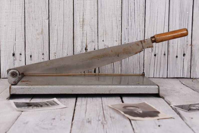 May include: Vintage metal paper cutter with a wooden handle. The blade is silver and shows signs of wear. The cutter is on a wooden surface, with old photographs scattered around.