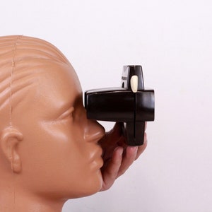 May include: A vintage, dark brown stereoscope viewer held up to a mannequin head. The viewer has a rectangular shape with a white button on top. The mannequin head is a light tan color.