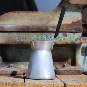May include: A silver coffee pot with a black handle sits on a wooden surface in front of a rusty metal vice.