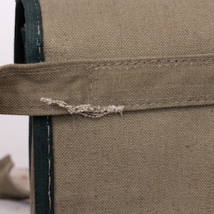 K&ouml;nnte beinhalten: Nahaufnahme einer olivgr&uuml;nen Canvas-Tasche mit dunkelgr&uuml;ner Paspelierung und Griff. Die Tasche hat einen silbernen Metallniet. Der Griff ist mit N&auml;hten befestigt und weist Abnutzungserscheinungen auf.