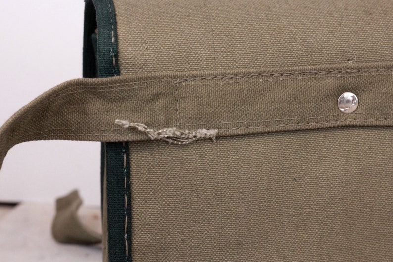 K&ouml;nnte beinhalten: Nahaufnahme einer olivgr&uuml;nen Canvas-Tasche mit dunkelgr&uuml;ner Paspelierung und Griff. Die Tasche hat einen silbernen Metallniet. Der Griff ist mit N&auml;hten befestigt und weist Abnutzungserscheinungen auf.