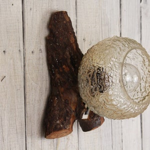 May include: A rustic wall sconce featuring a textured glass globe light fixture attached to a piece of natural wood. The wood is dark brown with a rough texture, and the light is a warm, inviting glow.