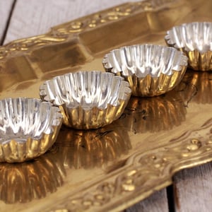 Pode incluir: Quatro pequenas formas de tarte caneladas com interiores prateados e exteriores dourados estão numa bandeja dourada e ornamentada. As formas estão dispostas em fila, refletindo a luz. A bandeja repousa sobre uma superfície de madeira clara e envelhecida.