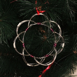 May include: A silver wire Christmas ornament with a circular design and pink beads. The ornament is hanging on a green Christmas tree.