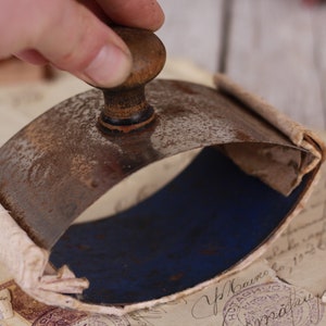 May include: An antique letter press with a wooden handle and a curved, metal base. The base has a blue interior and is surrounded by aged paper. The press is resting on a collection of old documents.