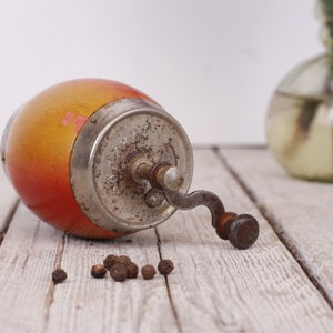 May include: An antique pepper grinder with a wooden body in shades of yellow and red, and a metal grinding mechanism. Several peppercorns are scattered nearby. The grinder is lying on a weathered wooden surface.