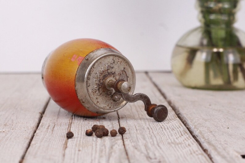 May include: An antique pepper grinder with a wooden body in shades of yellow and red, and a metal grinding mechanism. Several peppercorns are scattered nearby. The grinder is lying on a weathered wooden surface.