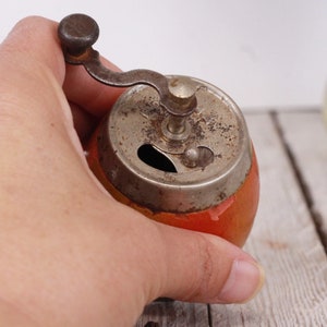 May include: A vintage pepper grinder with a red and orange painted body and a tarnished metal grinding mechanism. The grinder has a small crank handle and a round top. The pepper grinder is a kitchen utensil.