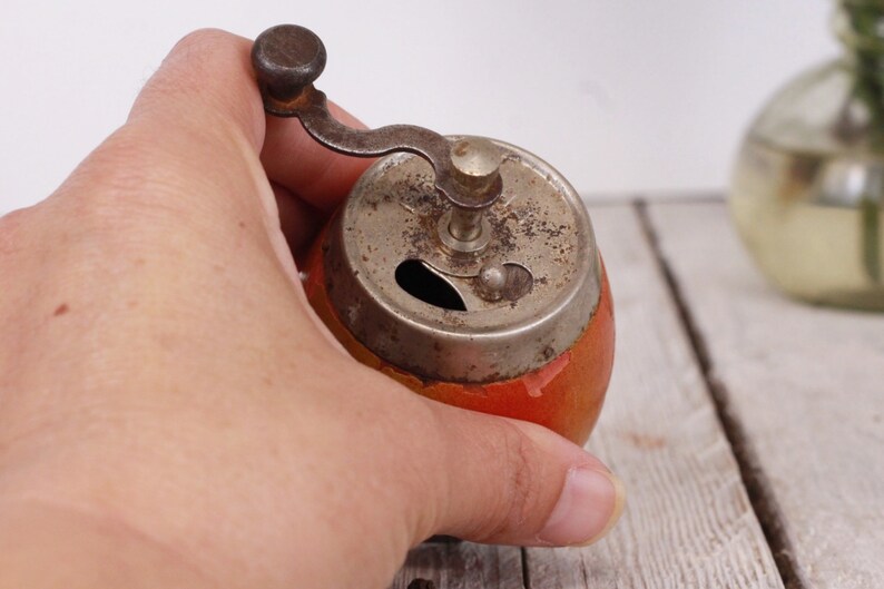 May include: A vintage pepper grinder with a red and orange painted body and a tarnished metal grinding mechanism. The grinder has a small crank handle and a round top. The pepper grinder is a kitchen utensil.
