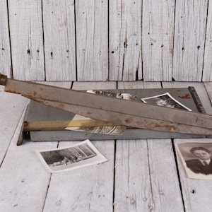 May include: An antique paper cutter with a wooden handle and a gray metal blade. The cutter is resting on a gray surface, with several black and white photographs scattered around it. The background is a weathered, white wooden surface.
