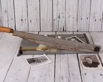 May include: An antique paper cutter with a wooden handle and a gray metal blade. The cutter is resting on a gray surface, with several black and white photographs scattered around it. The background is a weathered, white wooden surface.