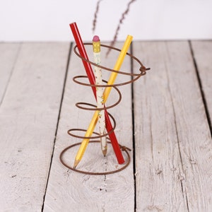 May include: A rustic, spiral-shaped metal pencil holder with three pencils. One pencil is red, one is yellow, and the third is white with a floral pattern. The holder sits on a white wooden surface.