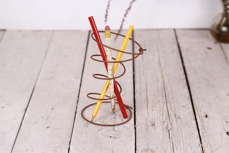 May include: A rustic, spiral-shaped metal pencil holder with three pencils. One pencil is red, one is yellow, and the third is white with a floral pattern. The holder sits on a white wooden surface.