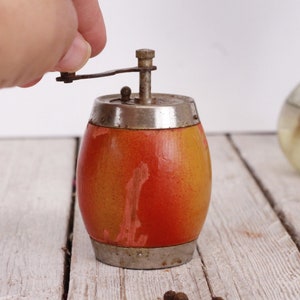 May include: A vintage pepper mill with a barrel-shaped body. The wooden body transitions from yellow to orange. It has a silver-colored top and bottom with a crank for grinding. A classic kitchen tool.