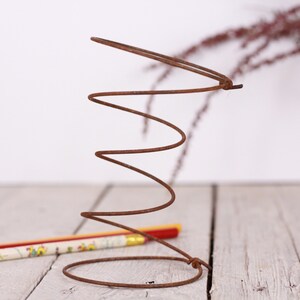 May include: A rusty brown metal spring stands upright, its coiled form prominent against a blurred background. A pencil with a floral design lies near the base of the spring. The image has a rustic, industrial feel.