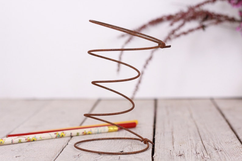 May include: A rusty brown metal spring stands upright, its coiled form prominent against a blurred background. A pencil with a floral design lies near the base of the spring. The image has a rustic, industrial feel.