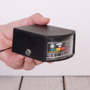May include: A black rectangular device with a curved, transparent front panel displaying a gauge with coloured indicators and numbers. The device has a small screw and a wire attached. The text on the gauge is in a foreign language.