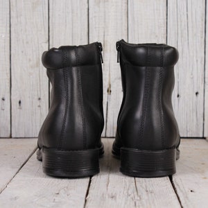 Peut inclure: Une paire de bottines en cuir noir avec un petit talon. Les bottines ont une fermeture &eacute;clair &agrave; l'arri&egrave;re et un col rembourr&eacute;. Les bottines sont pr&eacute;sent&eacute;es sur un fond en bois blanc.