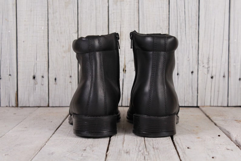 Peut inclure: Une paire de bottines en cuir noir avec un petit talon. Les bottines ont une fermeture &eacute;clair &agrave; l'arri&egrave;re et un col rembourr&eacute;. Les bottines sont pr&eacute;sent&eacute;es sur un fond en bois blanc.
