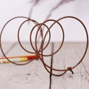 May include: A rusty brown metal wire sculpture with intertwined circular shapes. The sculpture is on a light gray wooden surface, with a colorful pencil in the background. The wire is bent into several loops and circles.