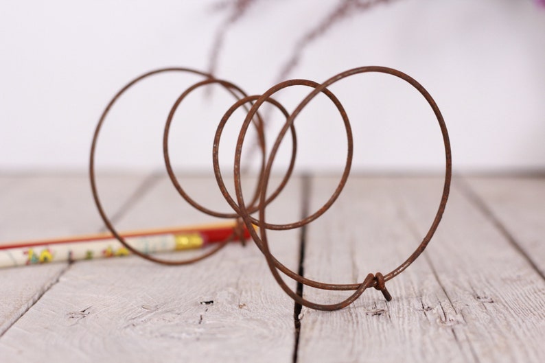 May include: A rusty brown metal wire sculpture with intertwined circular shapes. The sculpture is on a light gray wooden surface, with a colorful pencil in the background. The wire is bent into several loops and circles.