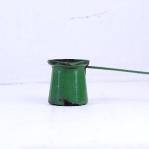 Puede incluir: Una cafetera de metal verde con un mango largo. La cafetera tiene un aspecto desgastado y vintage.