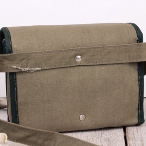 K&ouml;nnte beinhalten: Eine olivgr&uuml;ne Canvas-Tasche mit dunkelgr&uuml;ner Einfassung und passendem Schulterriemen. Die Tasche hat eine rechteckige Form mit Klappenverschluss und einem silbernen Druckknopf. Der Riemen ist mit einem silbernen Knopf an der Tasche befestigt.