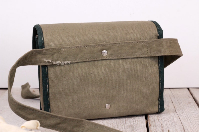 K&ouml;nnte beinhalten: Eine olivgr&uuml;ne Canvas-Tasche mit dunkelgr&uuml;ner Einfassung und passendem Schulterriemen. Die Tasche hat eine rechteckige Form mit Klappenverschluss und einem silbernen Druckknopf. Der Riemen ist mit einem silbernen Knopf an der Tasche befestigt.