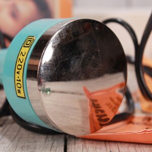 May include: A close-up of a vintage teal and silver beauty tool with a black cord. The tool has a label that reads "220V-10W". The background includes an orange paper and a blurred image.