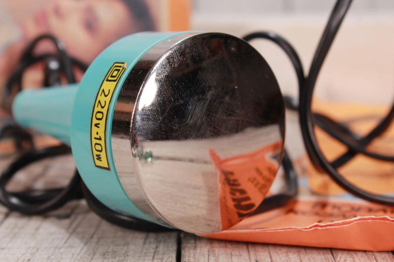May include: A close-up of a vintage teal and silver beauty tool with a black cord. The tool has a label that reads "220V-10W". The background includes an orange paper and a blurred image.