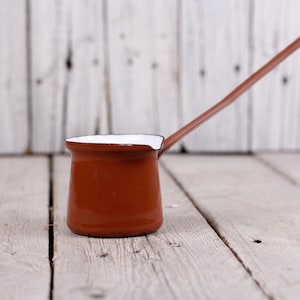 May include: A brown enamel coffee pot with a white interior and a long handle. The pot is sitting on a wooden surface.
