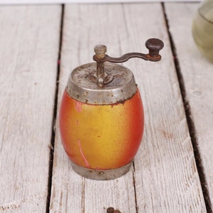 May include: An antique pepper grinder with a wooden barrel-shaped body, painted in shades of yellow and red. It has a metal top, bottom, and a crank handle. The grinder sits on a weathered, white wooden surface.
