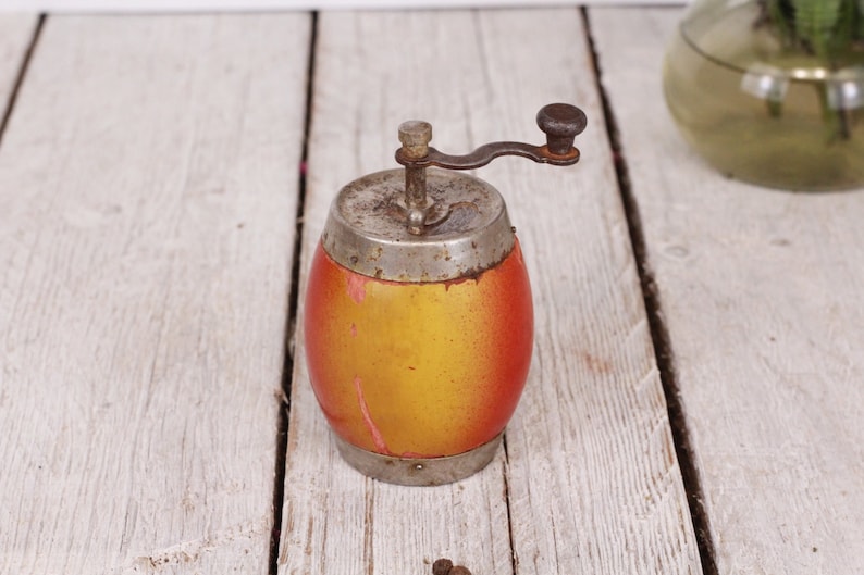 May include: An antique pepper grinder with a wooden barrel-shaped body, painted in shades of yellow and red. It has a metal top, bottom, and a crank handle. The grinder sits on a weathered, white wooden surface.