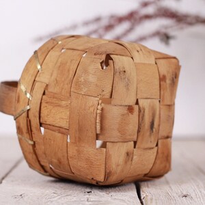 Puede incluir: Una peque&ntilde;a cesta tejida con asa. La cesta est&aacute; hecha de tiras de madera marr&oacute;n claro, tejidas en un patr&oacute;n cuadrado. El asa es de madera maciza. La cesta est&aacute; sobre una superficie de madera blanca.