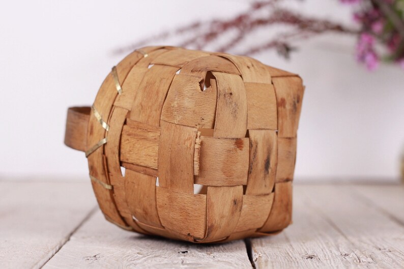 Puede incluir: Una peque&ntilde;a cesta tejida con asa. La cesta est&aacute; hecha de tiras de madera marr&oacute;n claro, tejidas en un patr&oacute;n cuadrado. El asa es de madera maciza. La cesta est&aacute; sobre una superficie de madera blanca.