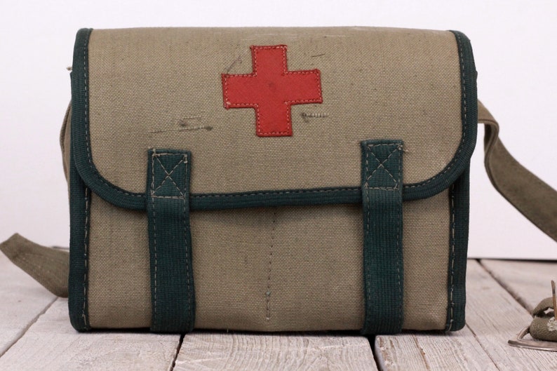 K&ouml;nnte beinhalten: Vintage-Sanit&auml;tskasten aus olivgr&uuml;nem Canvas. Er hat ein rotes Kreuz, dunkelgr&uuml;ne Verzierungen und Riemen. Die Tasche hat eine rechteckige Form mit Klappe und Schulterriemen.