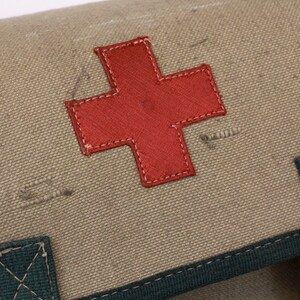 K&ouml;nnte beinhalten: Nahaufnahme einer Erste-Hilfe-Tasche aus Segeltuch mit einem markanten roten Kreuz. Die Tasche ist in einem ged&auml;mpften Olivgr&uuml;n gehalten und hat dunkelgr&uuml;ne Verzierungen und Riemen. Das rote Kreuz ist auf die Tasche gen&auml;ht.