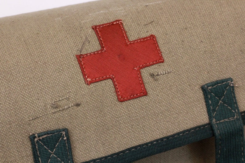 K&ouml;nnte beinhalten: Nahaufnahme einer Erste-Hilfe-Tasche aus Segeltuch mit einem markanten roten Kreuz. Die Tasche ist in einem ged&auml;mpften Olivgr&uuml;n gehalten und hat dunkelgr&uuml;ne Verzierungen und Riemen. Das rote Kreuz ist auf die Tasche gen&auml;ht.