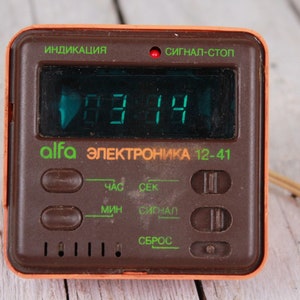 May include: A retro brown and orange digital alarm clock showing 03:14 in green. The clock face features the text "alfa ЭЛЕКТРОНИКА 12-41" and various buttons and switches for setting the time and alarm.
