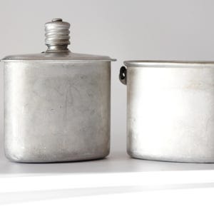 Puede incluir: Dos cantimploras metálicas vintage. La cantimplora de la izquierda tiene una tapa de rosca y forma rectangular. La cantimplora de la derecha tiene un asa y una forma rectangular similar. Ambas son de color plateado y parecen ser de aluminio.