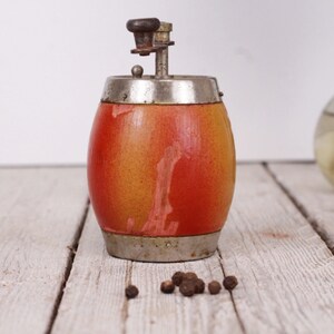 May include: A vintage pepper grinder with a barrel-shaped body. The wooden body transitions from yellow to red, with metal bands at the top and bottom. A small pile of peppercorns sits nearby. A decorative item for the kitchen.