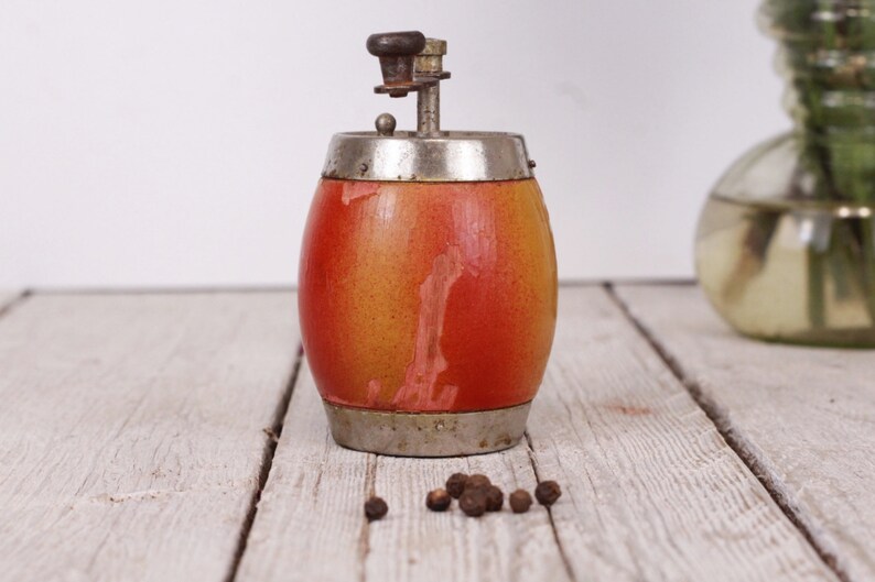 May include: A vintage pepper grinder with a barrel-shaped body. The wooden body transitions from yellow to red, with metal bands at the top and bottom. A small pile of peppercorns sits nearby. A decorative item for the kitchen.