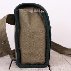 K&ouml;nnte beinhalten: Olivgr&uuml;ne Messenger Bag aus Canvas mit dunkelgr&uuml;ner Einfassung und einem langen, verstellbaren Riemen. Die Tasche hat eine rechteckige Form mit einem Klappenverschluss und einer Metallschnalle. Die Tasche ist aus strapazierf&auml;higem Canvas-Material.