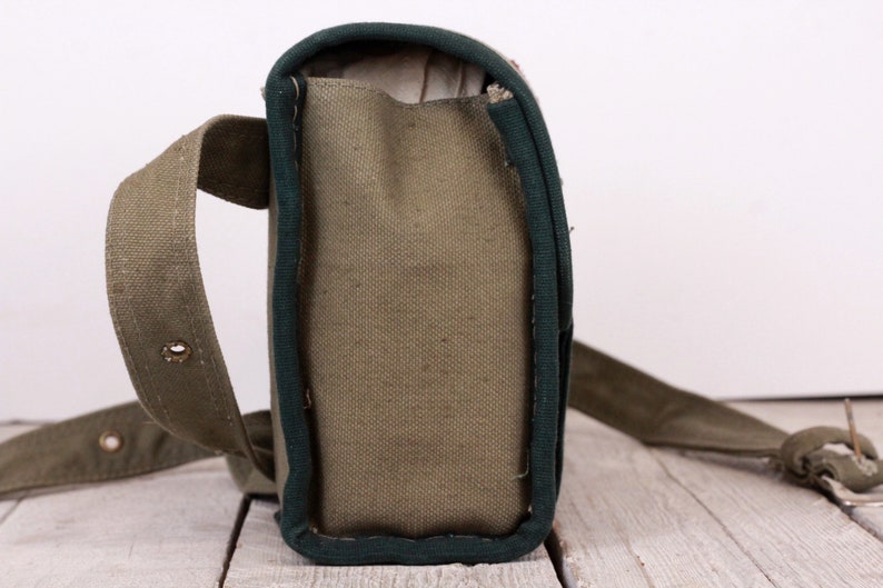 K&ouml;nnte beinhalten: Olivgr&uuml;ne Messenger Bag aus Canvas mit dunkelgr&uuml;ner Einfassung und einem langen, verstellbaren Riemen. Die Tasche hat eine rechteckige Form mit einem Klappenverschluss und einer Metallschnalle. Die Tasche ist aus strapazierf&auml;higem Canvas-Material.