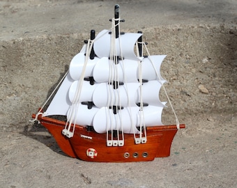 May include: A small wooden toy sailboat with white sails and black accents. The sailboat is sitting on a gray concrete surface.