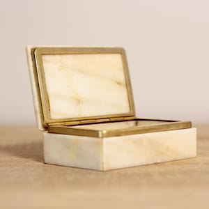 May include: A small, rectangular box made of white marble with a brass hinge. The box is open, revealing the interior, which is also made of white marble.